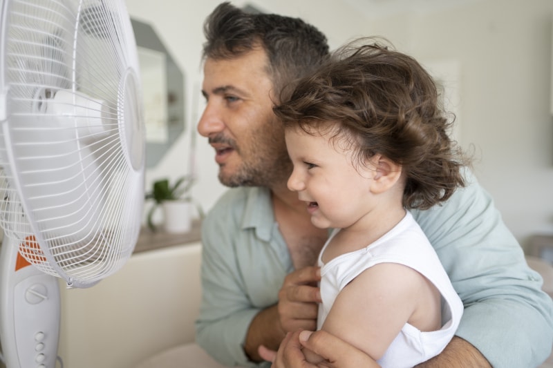 Father child is front of electric fan on hot summer day.